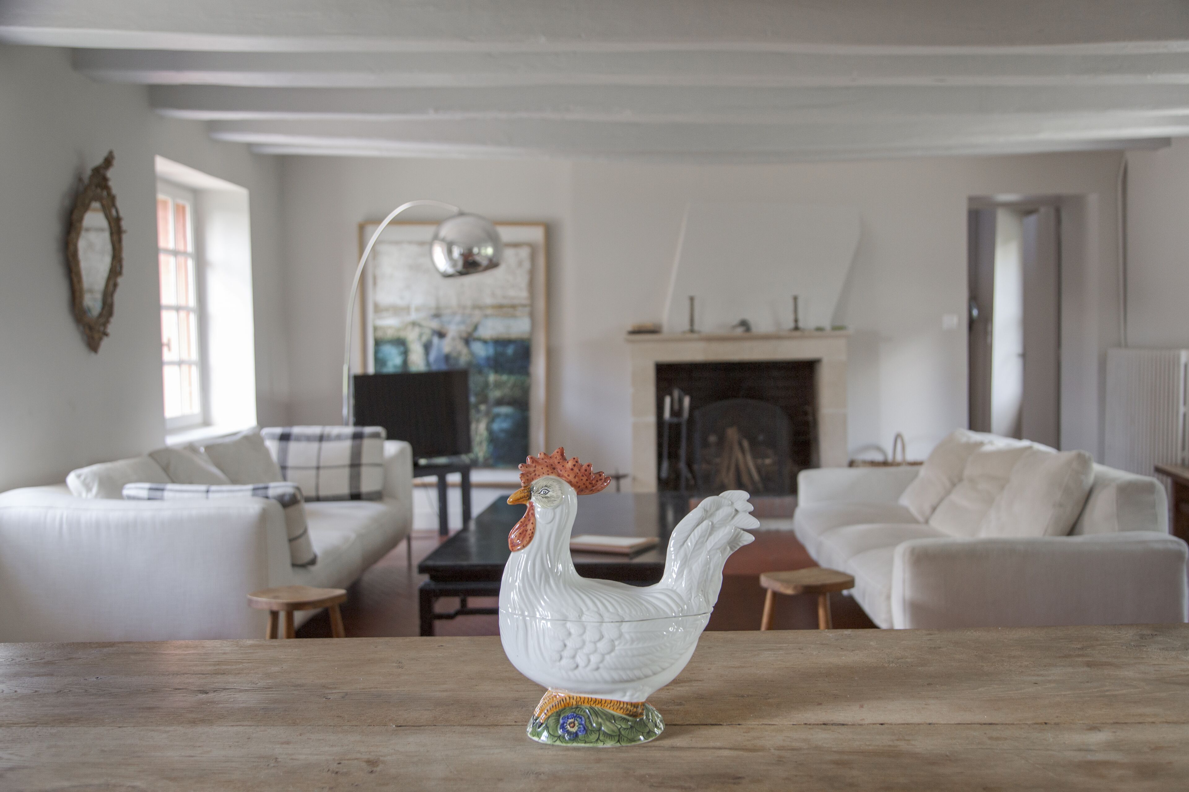 A white ceramic rooster on a wooden table in a living room with white sofas, a fireplace, and a framed painting on the wall.