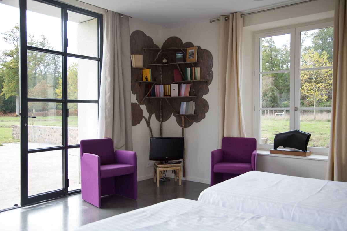 Une chambre d’hôtel moderne avec un grand lit, deux fauteuils violets, un téléviseur posé sur un meuble en bois et une bibliothèque adossée au mur.