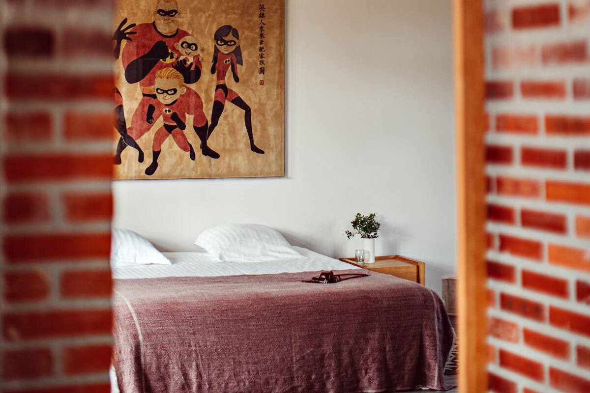 Une pièce avec un mur en briques, un plafond en bois, un lit recouvert d’une couverture rouge, une suspension rouge et un tableau encadré accroché au mur.