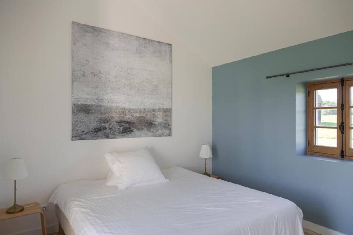 Une chambre à coucher comprenant un lit, une table de chevet, une lampe, une fenêtre avec des rideaux et une œuvre d’art accrochée au mur.