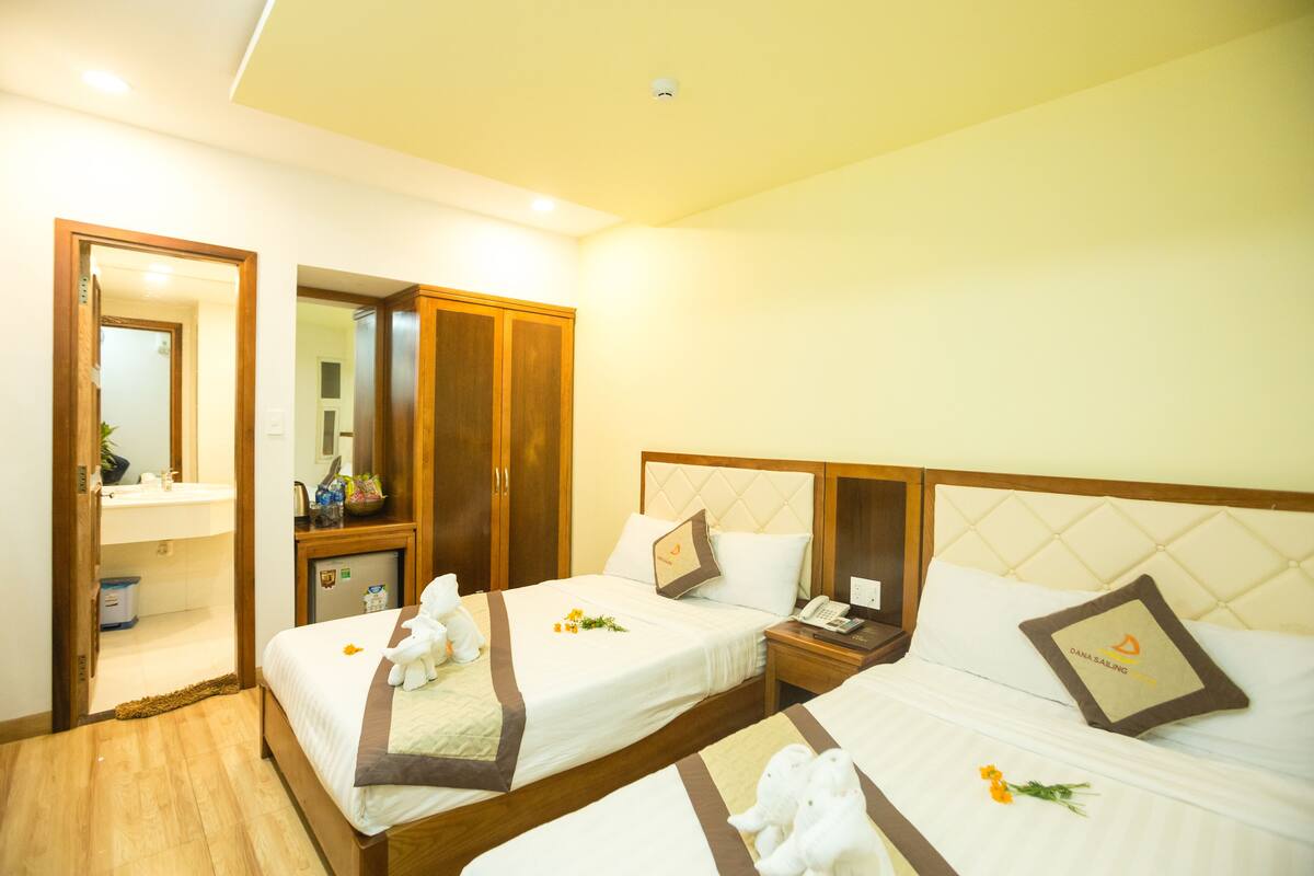 A hotel room with two beds, a wooden wardrobe, a bathroom with a mirror, and a small desk with a telephone.