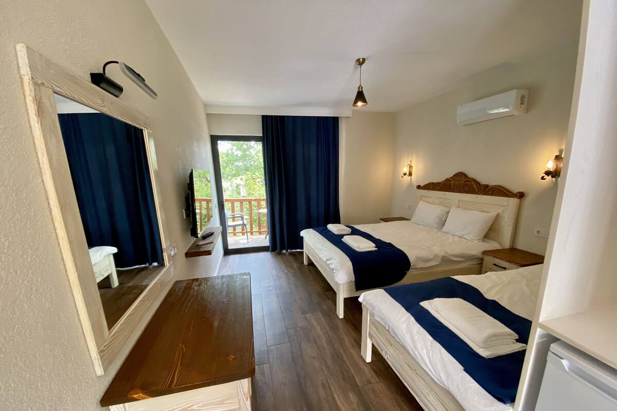 A hotel room with two beds, a wooden desk, a mirror, and a balcony with a view of greenery.