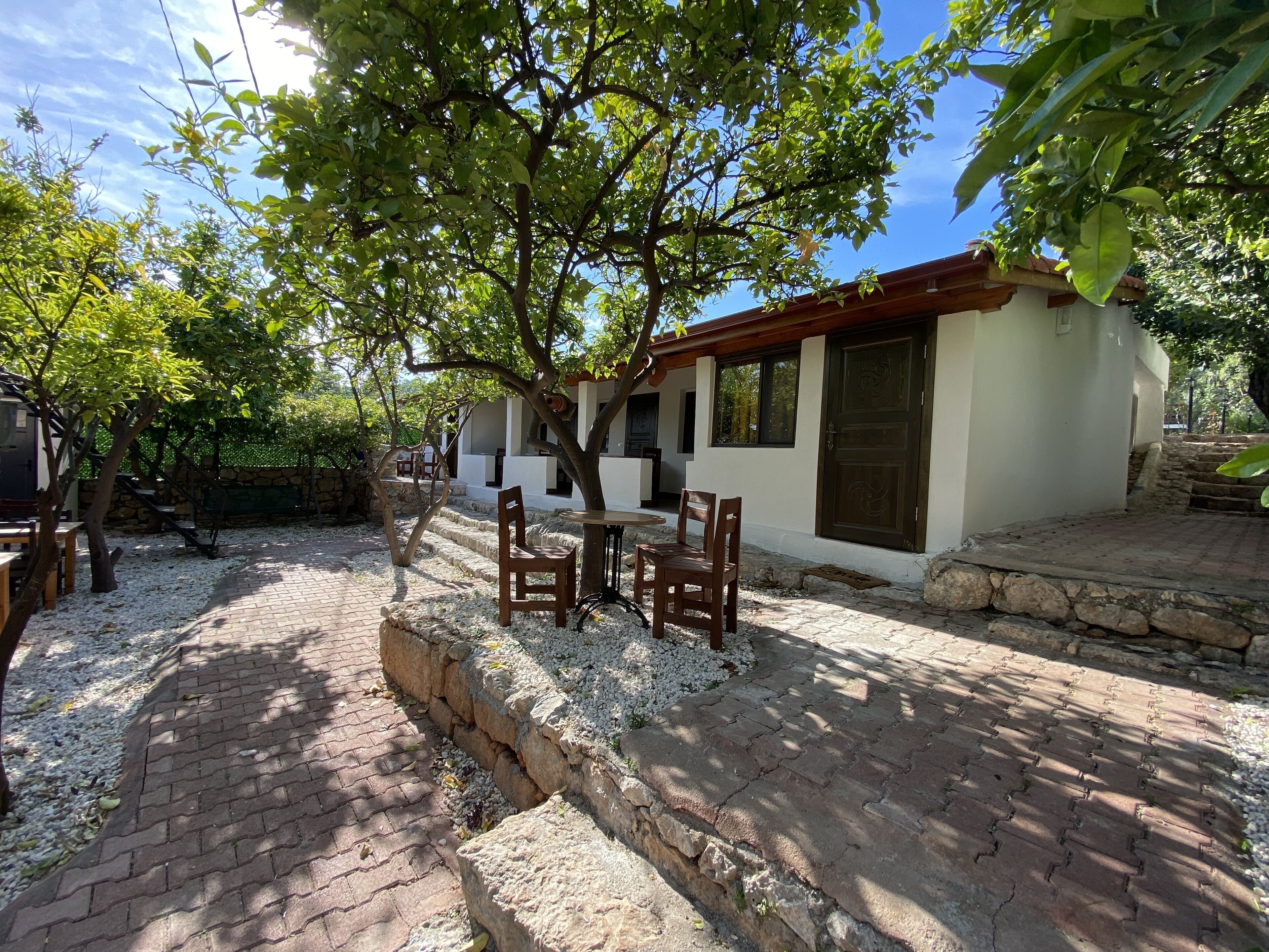 A patio area with a table and chairs, surrounded by trees and a white building with a brown door.