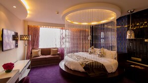 A modern bedroom with a circular bed, a hanging light fixture, and a view of the city through large windows.