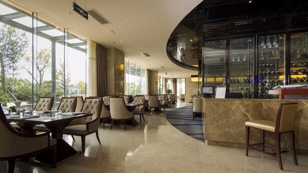 A modern hotel lobby with a curved reception desk, a bar area with glass display cases, and a dining area with round tables and chairs.