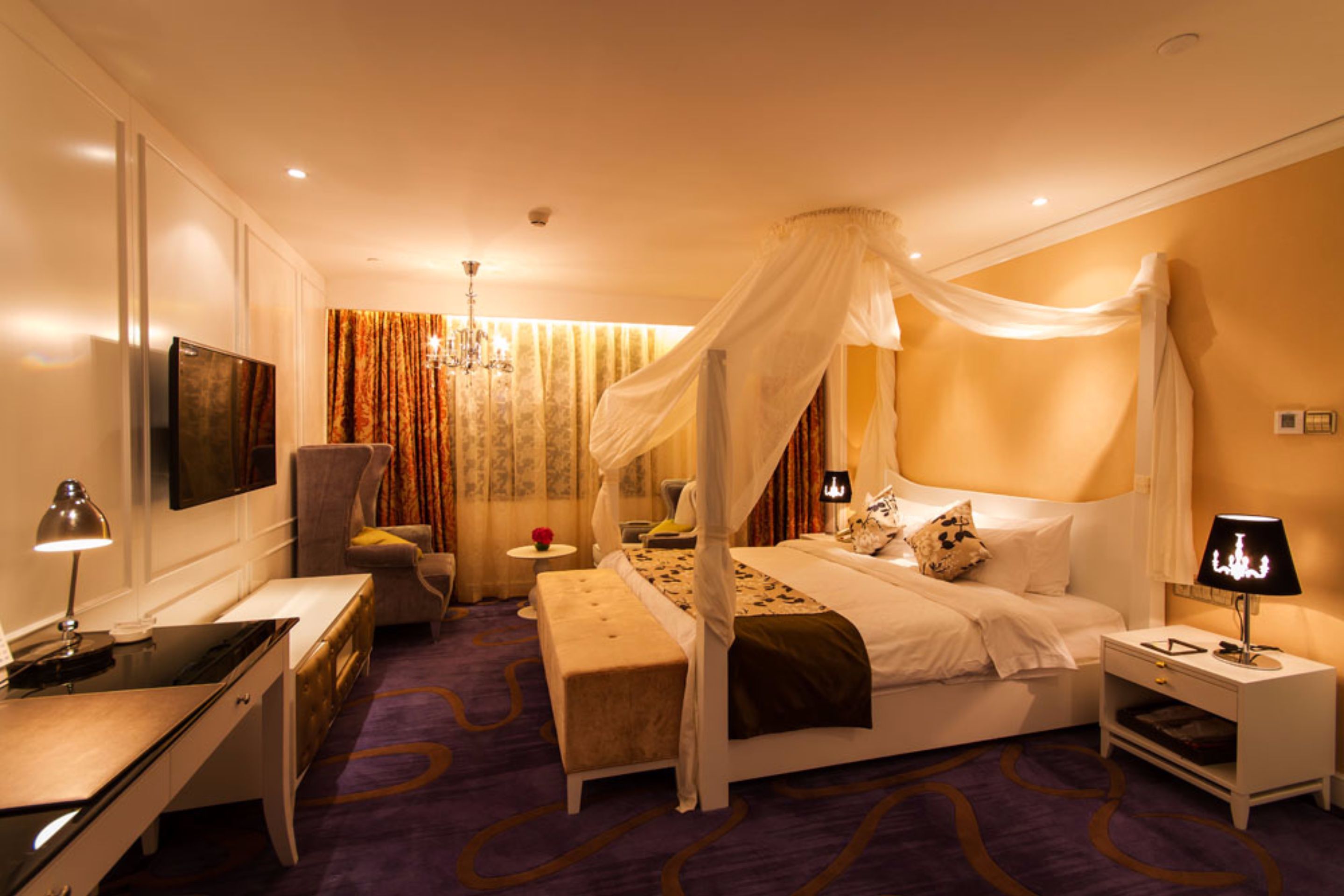 A bedroom with a four-poster bed, a desk, a chair, a bedside table, and a lamp.