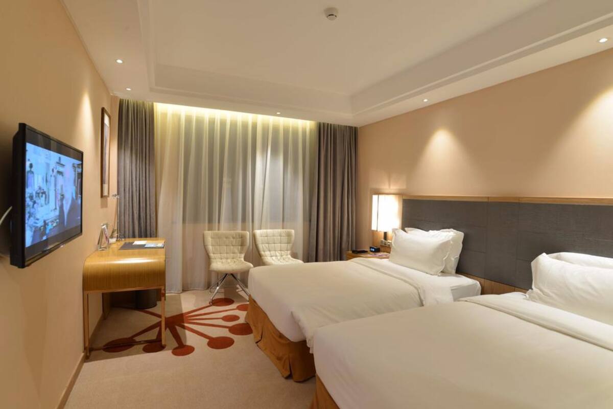 Une chambre d’hôtel avec deux lits, un bureau, une chaise, une télévision et une grande fenêtre avec des rideaux.