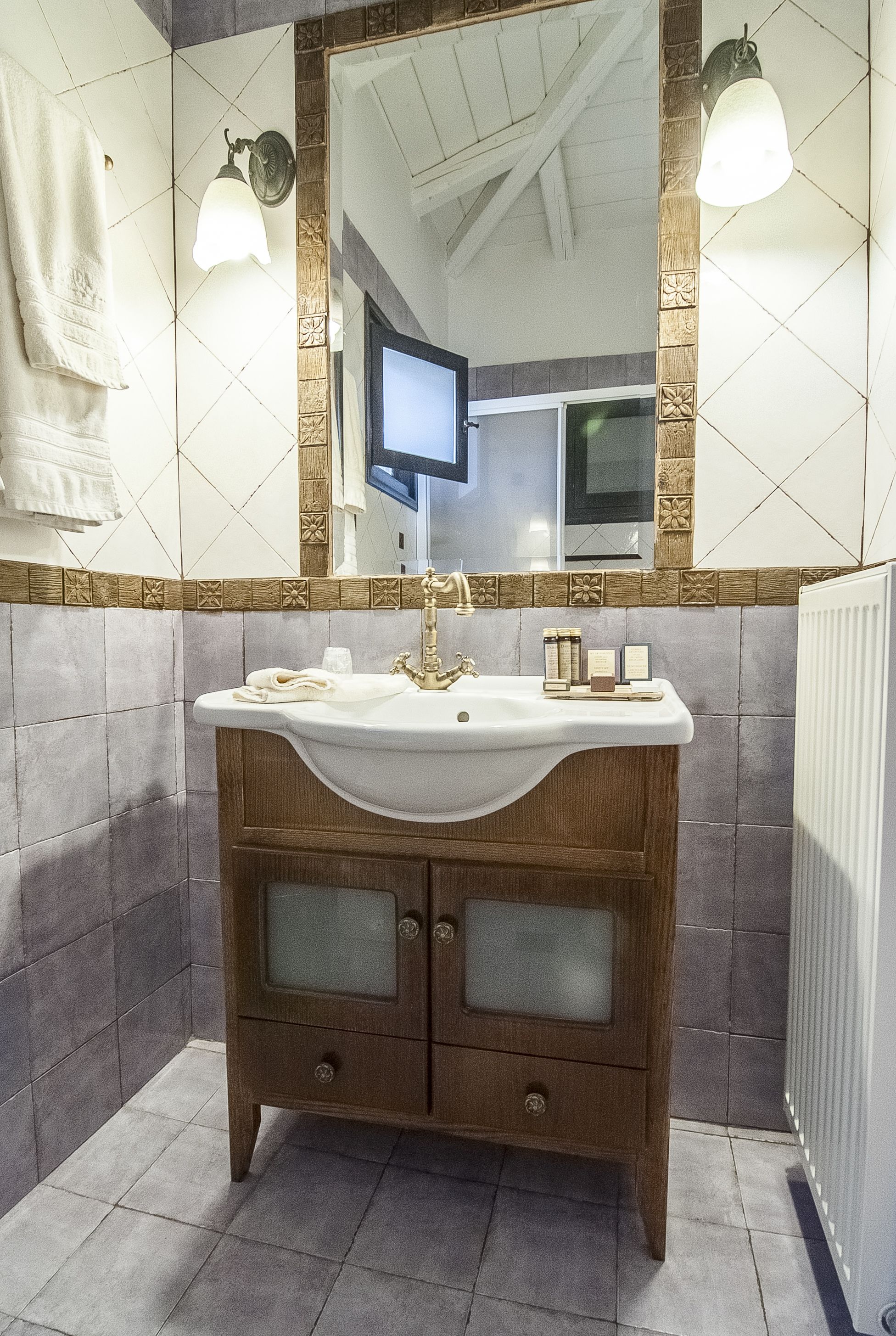 Un baño con un tocador de madera, un lavabo blanco y un espejo con marco decorativo. Hay dos luces fijadas a la pared y una toalla colgada.