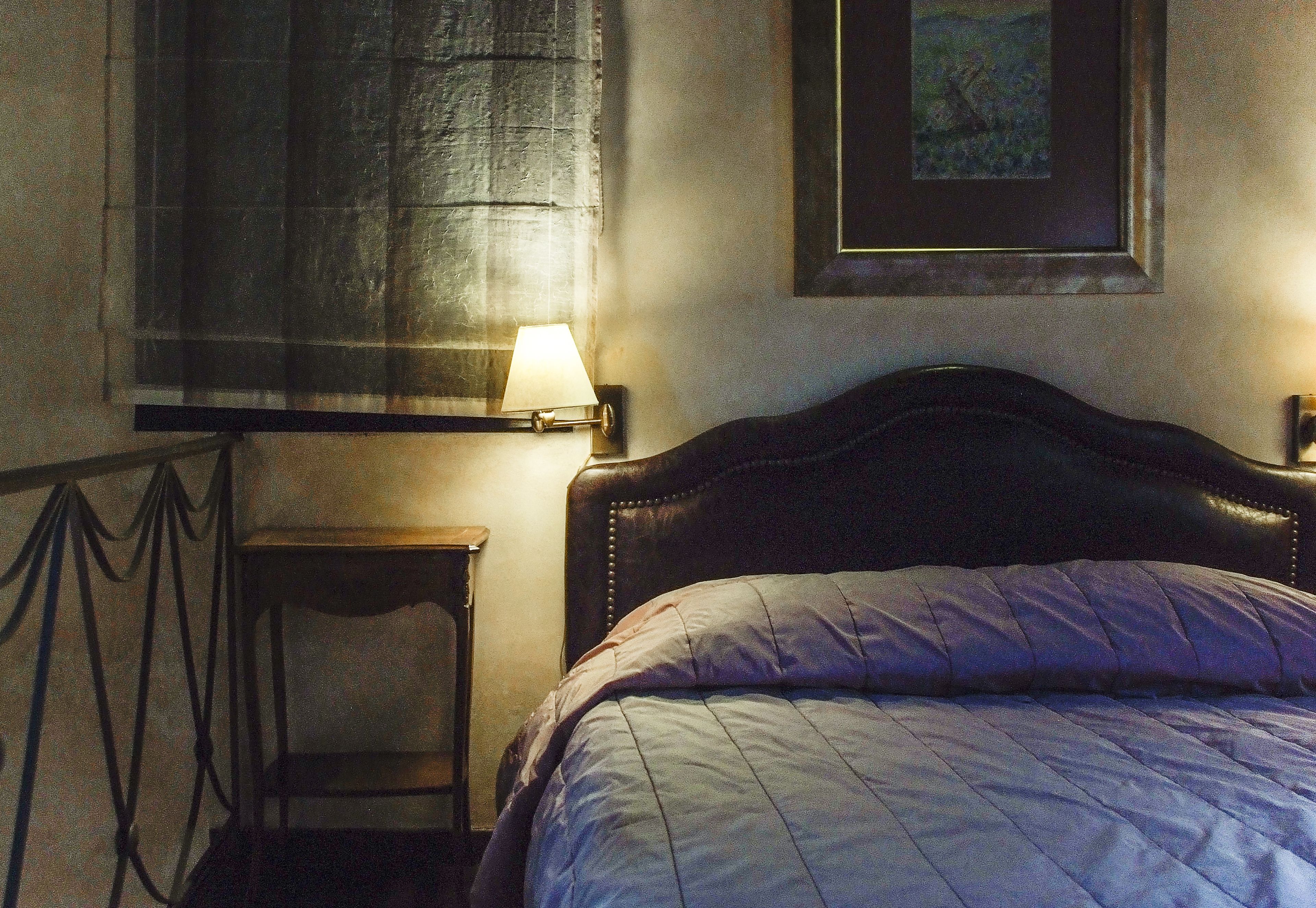 Quarto com cama, mesa de cabeceira, abajur, janela com cortina e um quadro na parede.