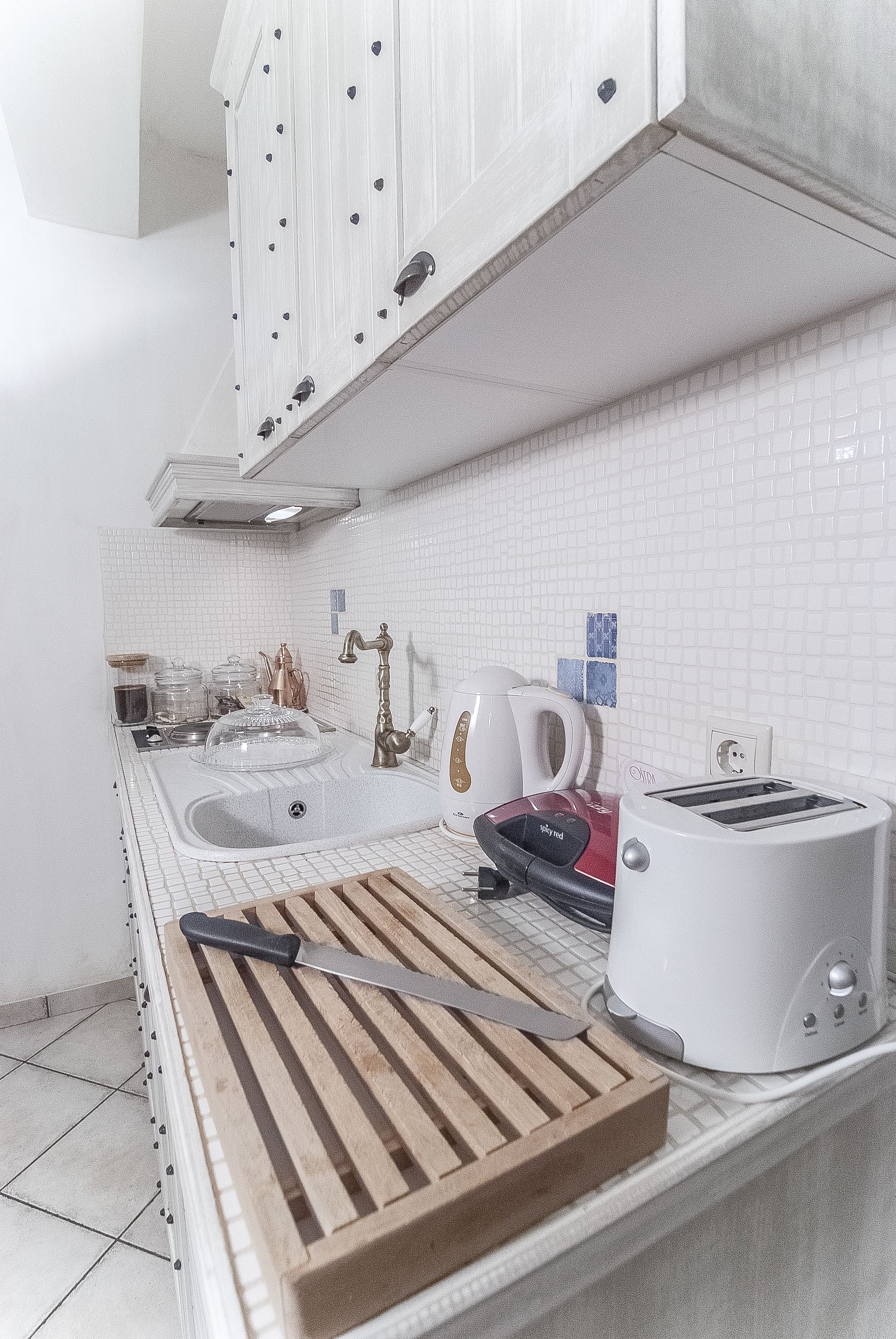 Una cocina con paredes de azulejos blancos, una tabla de cortar de madera, un cuchillo, un tostador y una tetera.