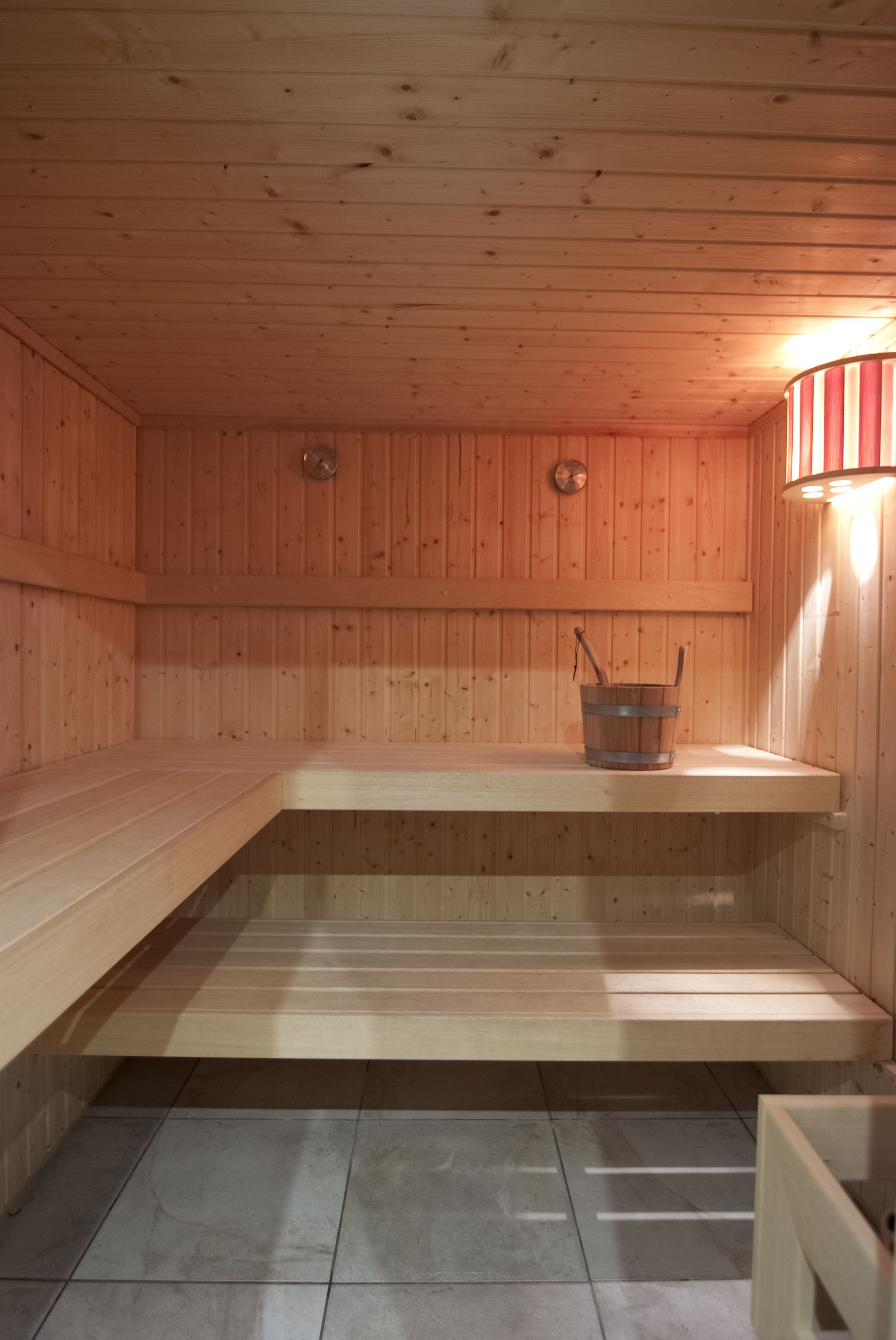 Uma sauna com paredes e bancos de madeira, um balde com uma escova e uma luminária fixada na parede.