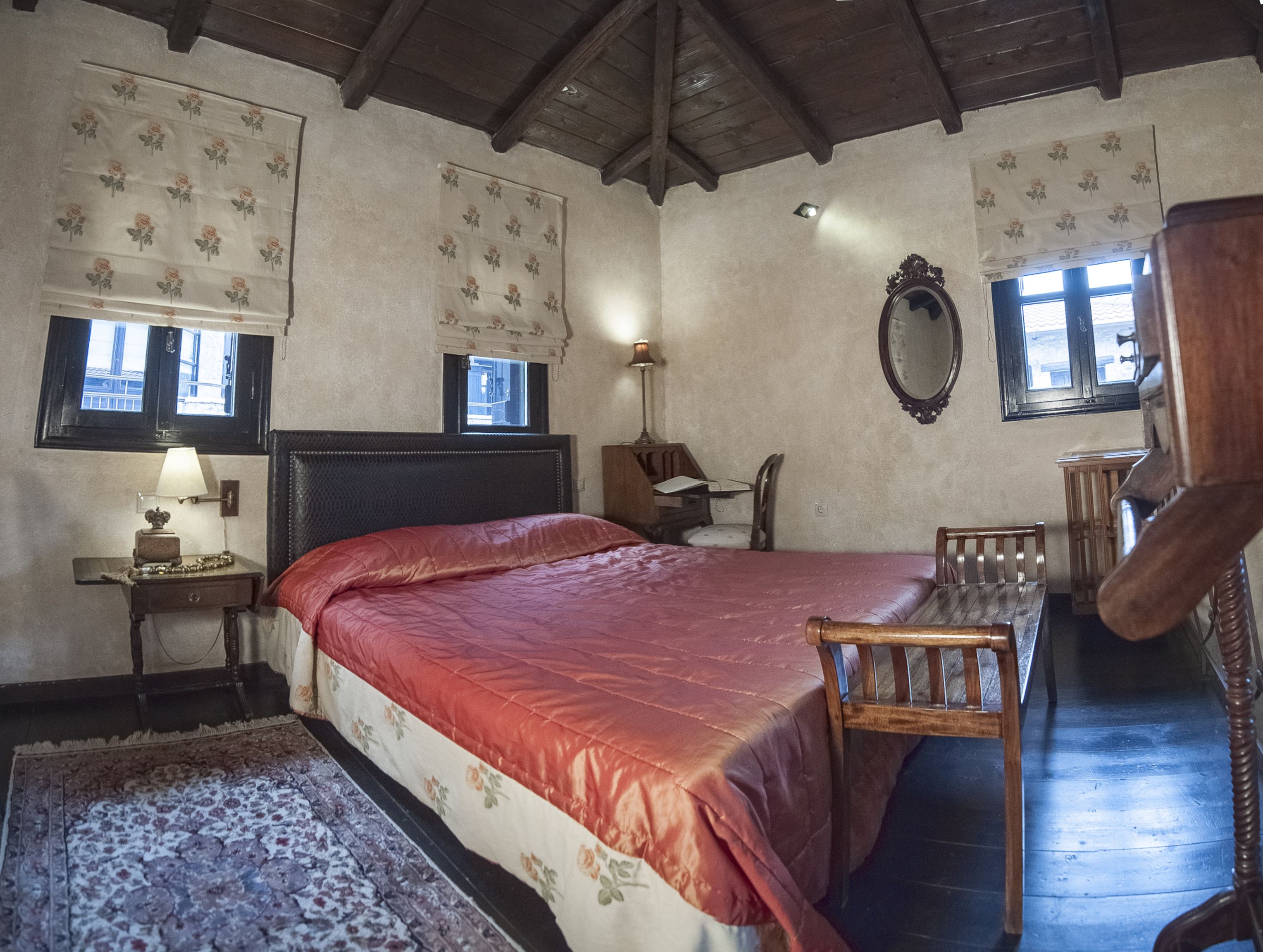 A bedroom with a large bed, a wooden desk, a chair, a mirror, and a window with floral curtains.