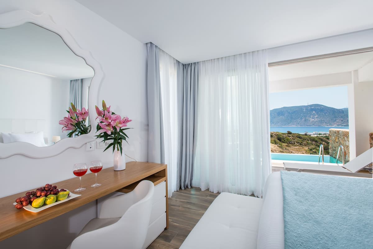 Une chambre d’hôtel moderne avec un bureau en bois, un lit, une grande fenêtre donnant sur la mer et les collines, ainsi qu’une table à manger avec des fruits et des verres.
