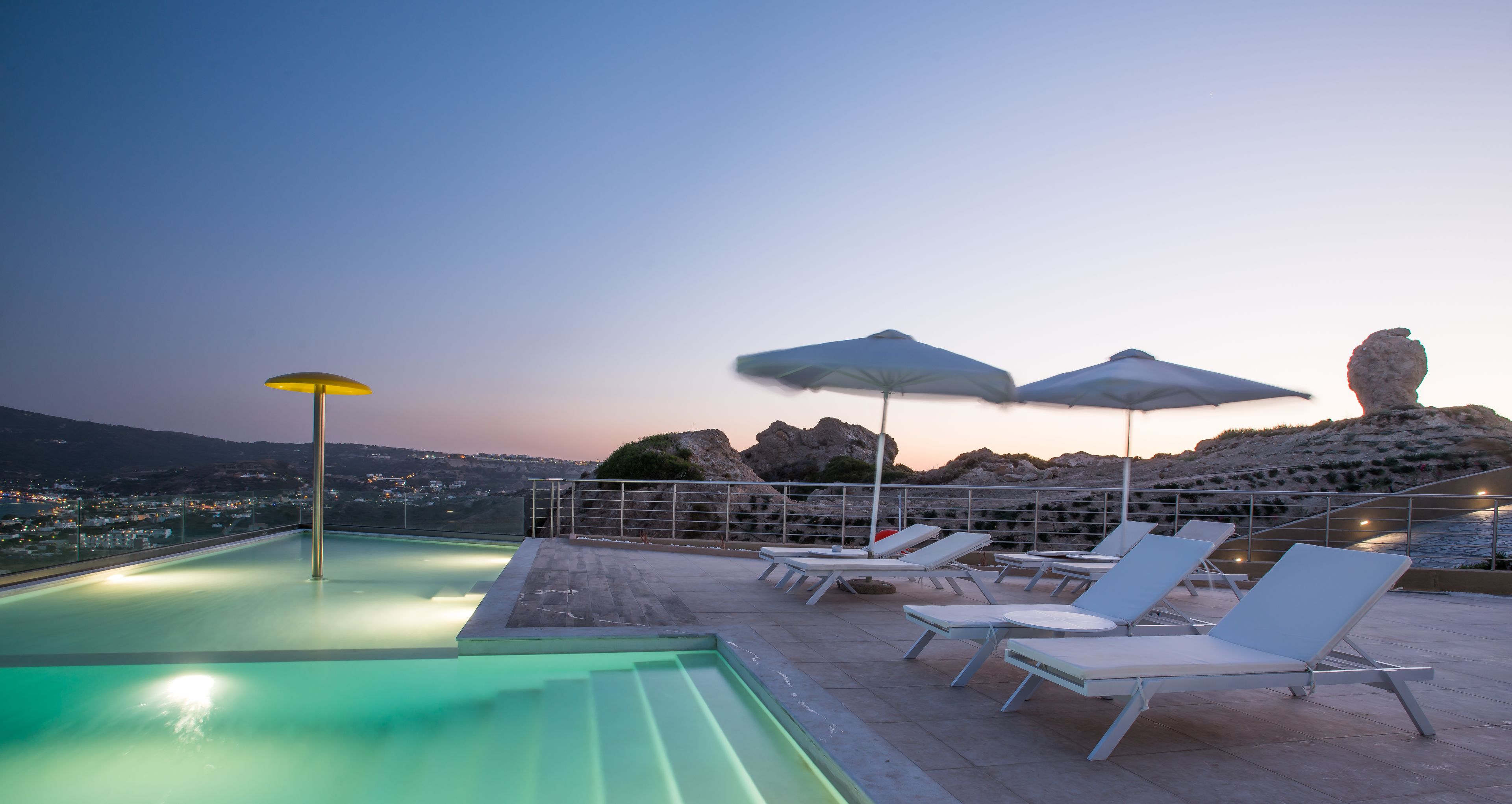 Area piscina sul tetto con lettini e ombrelloni, con vista su un paesaggio roccioso e una città in lontananza.