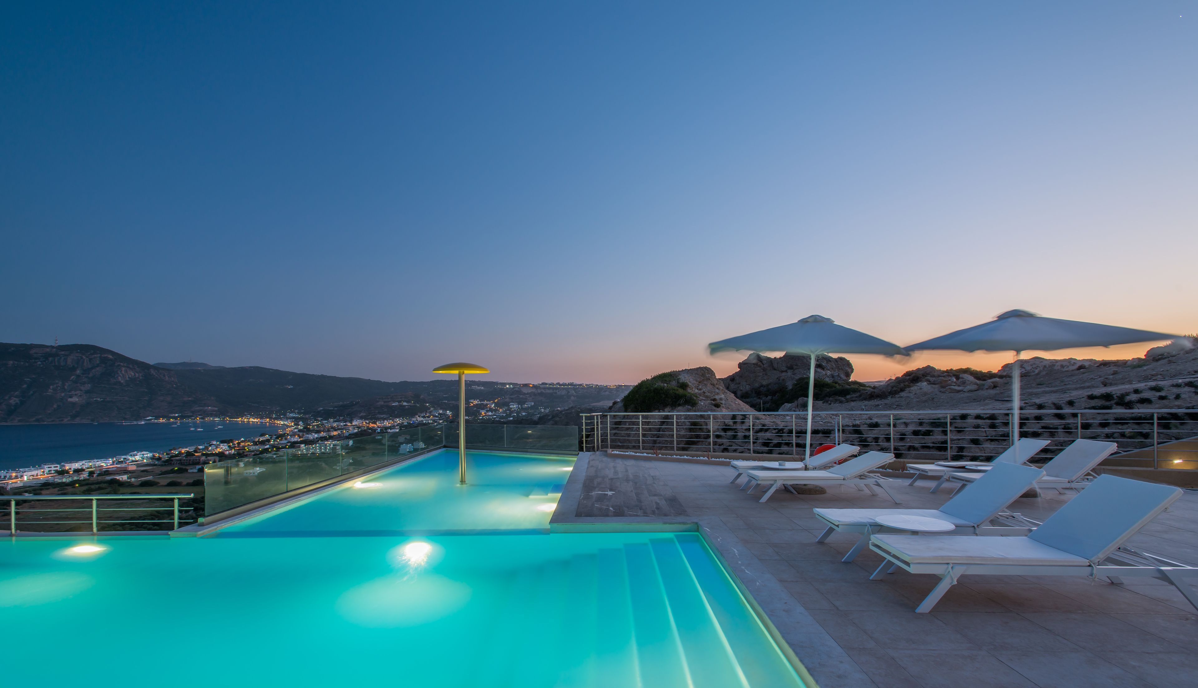 A rooftop infinity pool with lounge chairs and umbrellas, overlooking a coastal town and mountains at dusk.