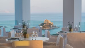 Un restaurant amb taules rodones, cadires blanques i vasos de vi. Es veu el mar i una illa rocosa al fons.