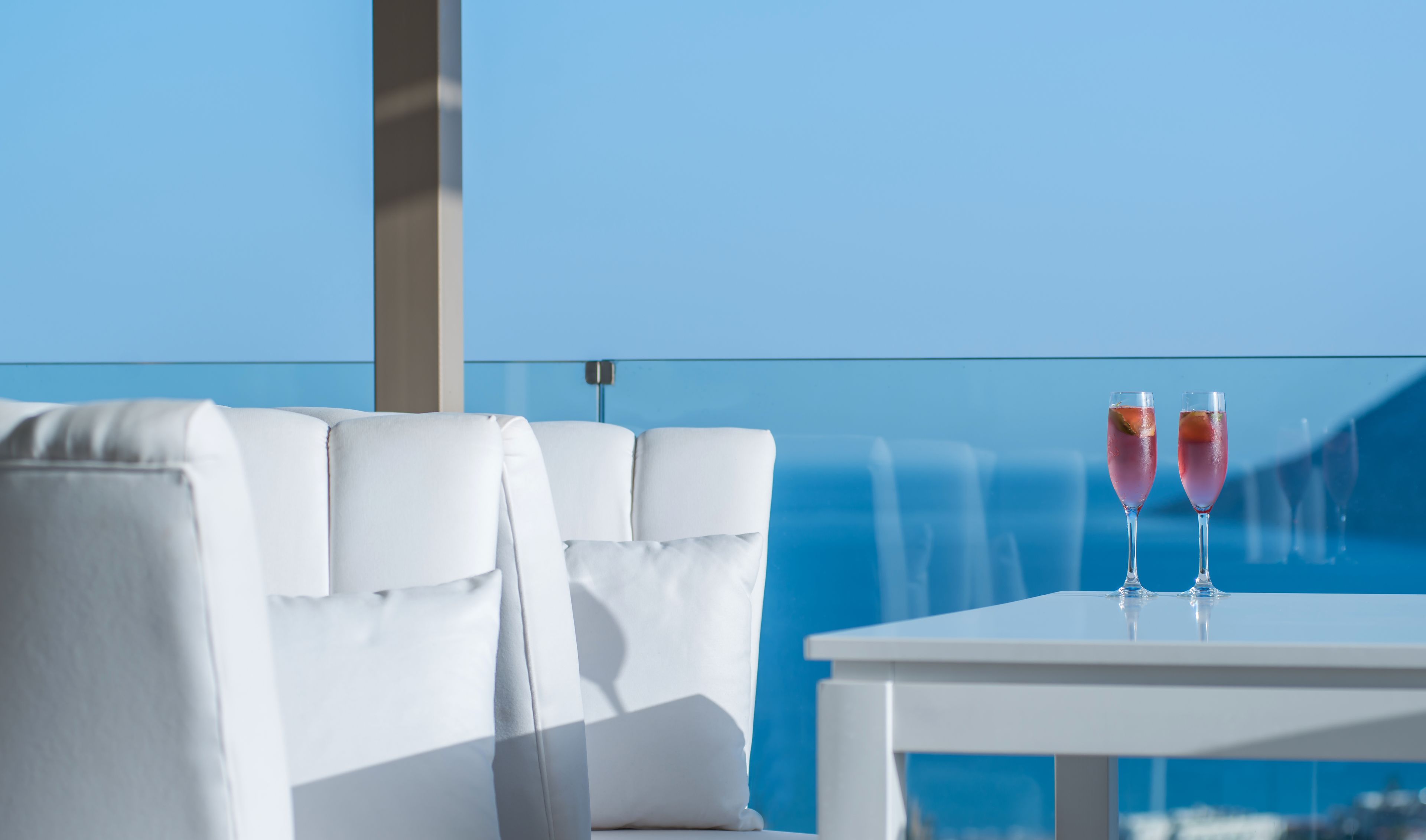 A balcony with white seating and two glasses of pink drink on a table, overlooking a clear blue sky.
