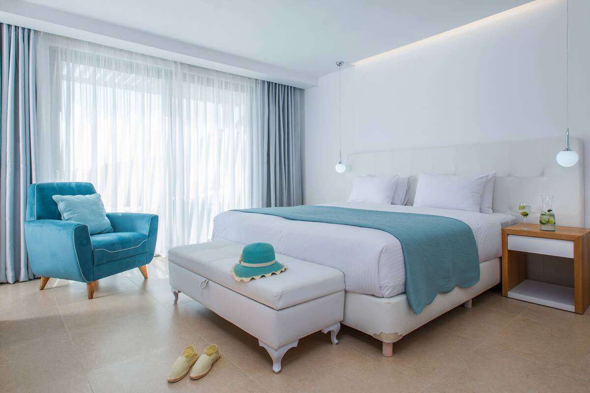 Une chambre à coucher moderne avec un grand lit, un fauteuil bleu, une table de chevet blanche et une fenêtre ornée de rideaux légers.