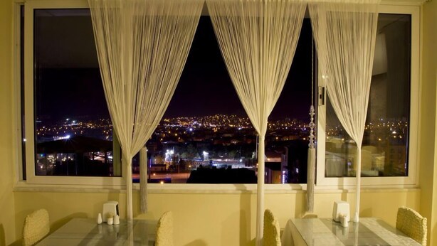 A dining area with a city view at night, featuring a table and chairs, a window with curtains, and a table setting with a vase and napkins. - Aygoren Otel Denizli (Denizli)