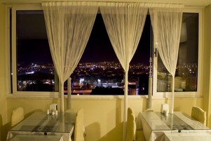 A dining area with a city view at night, featuring a table and chairs, a window with curtains, and a table setting with a vase and napkins.