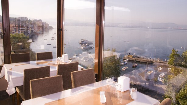 Un comedor con mesas y sillas, amplias ventanas que ofrecen vista a un frente marítimo con barcos y un paisaje urbano.