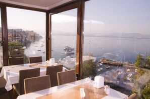 A dining area with tables and chairs, large windows offering a view of a waterfront with boats and a cityscape. - Kerimbey Otel (Gemlik)