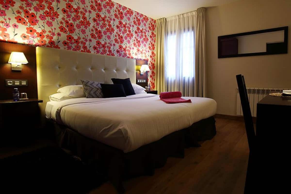 A hotel room with a large bed, two bedside lamps, a wooden floor, and a floral wallpaper design.