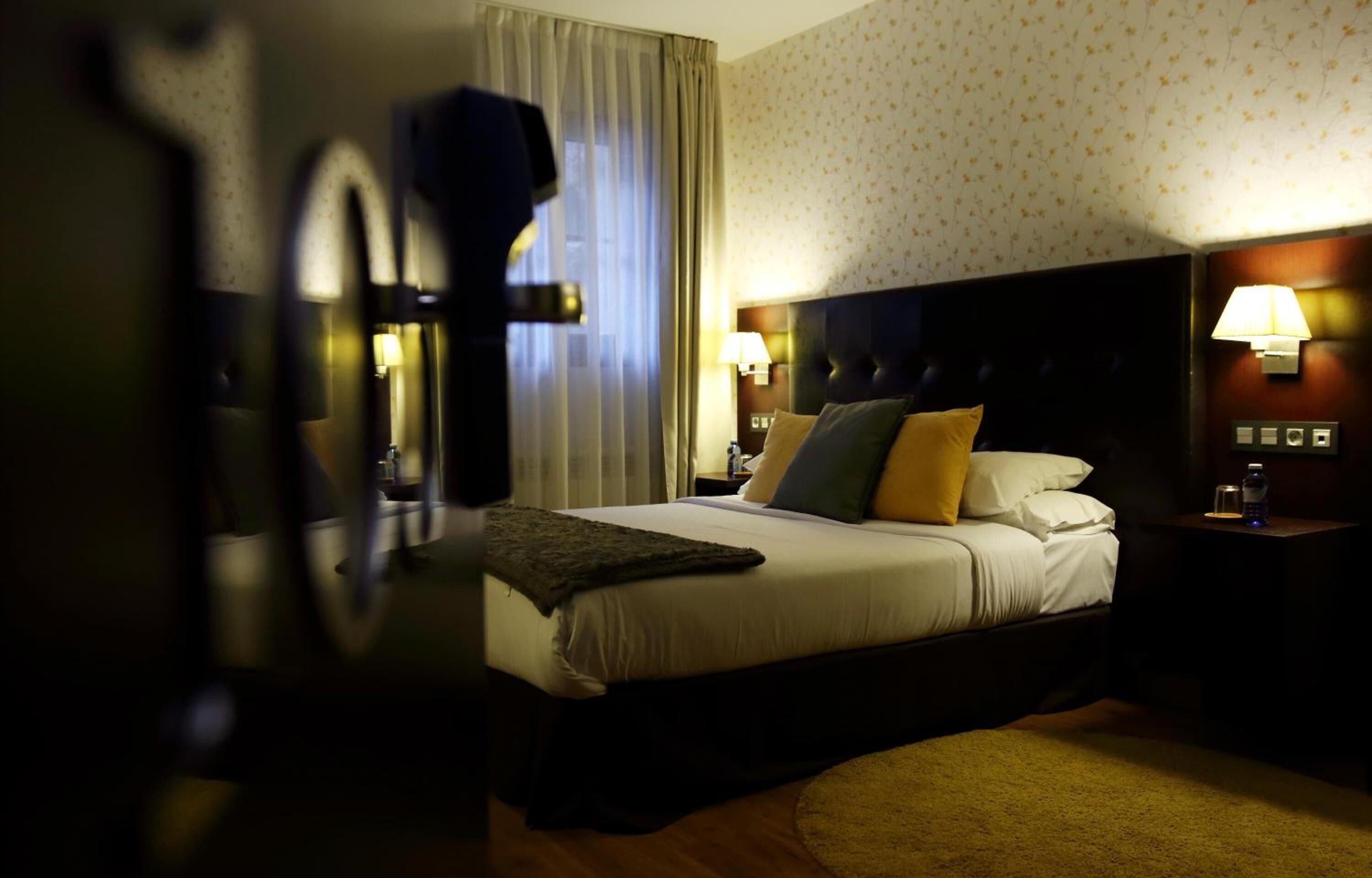 A hotel room with a large bed, two bedside lamps, a headboard, and a window with curtains.