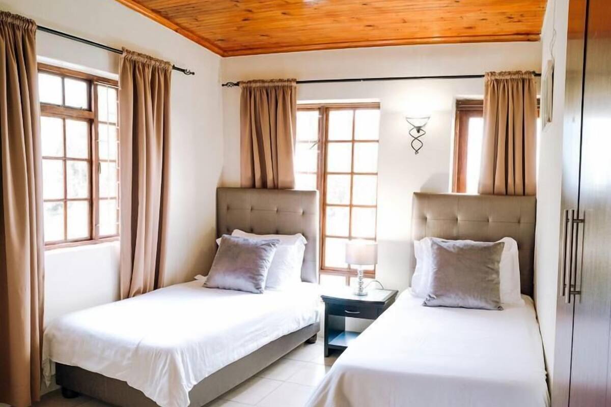 A room with two beds, wooden ceiling, and large windows with curtains.