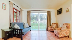 A living room with a sofa, armchairs, a TV, and a sliding glass door leading to a garden. - Kingfisher Hollow Exclusive Guest House (Cape Town)