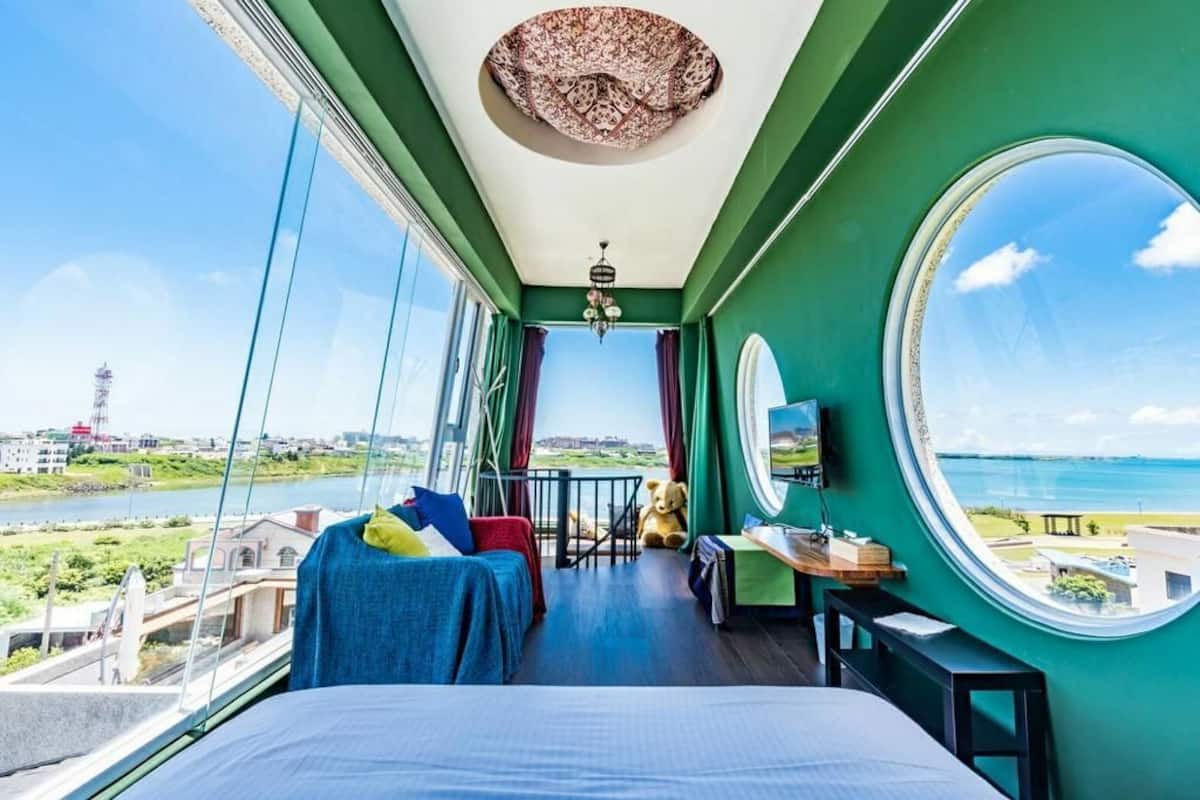 Habitación con balcón, cama, escritorio y un amplio ventanal con vistas al agua y al paisaje urbano.
