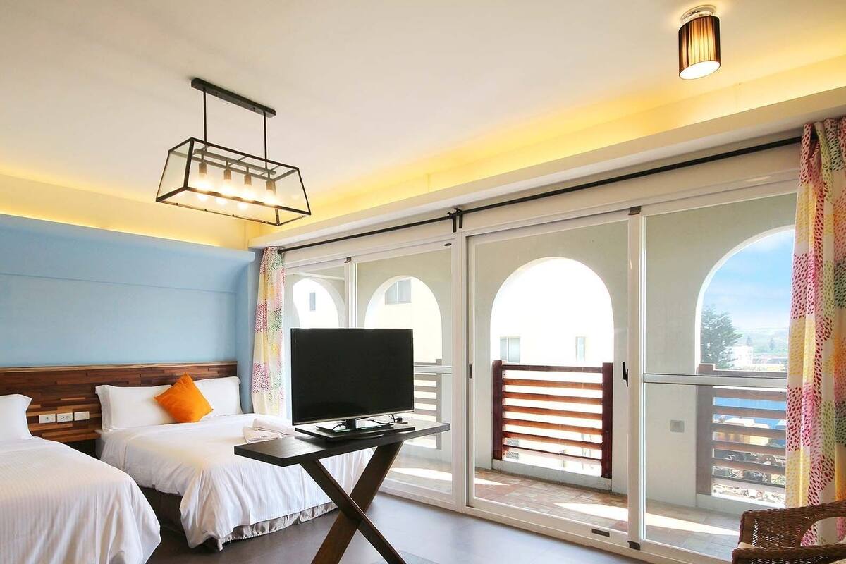 Habitación de hotel con dos camas, televisión y balcón con ventanales de arco.