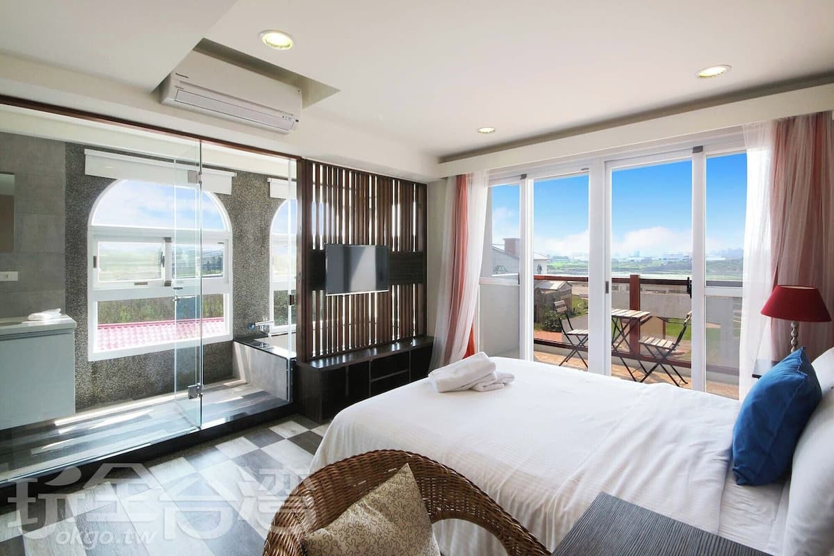 Habitación de hotel con una cama grande, televisión, balcón con vistas y zona de ducha de vidrio.