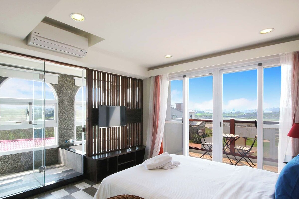A hotel room with a large bed, a TV, a balcony with a view, and a glass shower area.