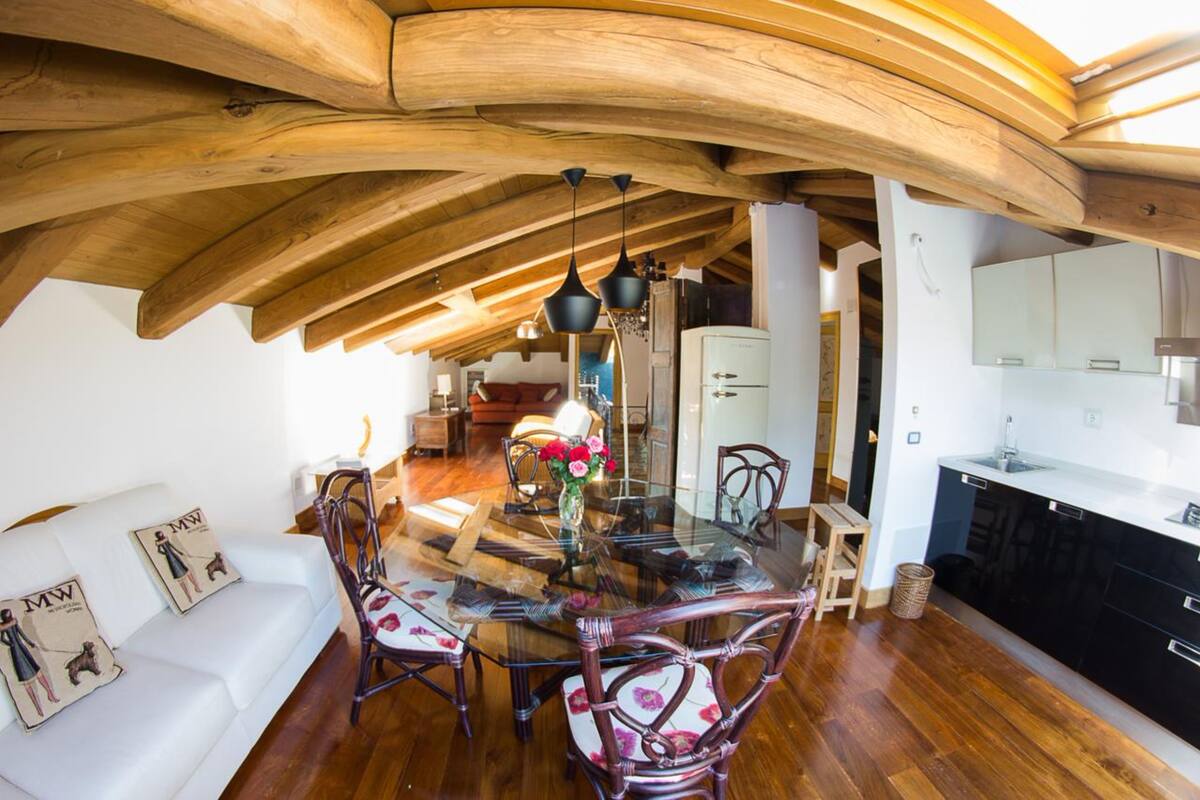 Un espace repas avec une table et des chaises en verre, des poutres en bois au plafond et un canapé blanc agrémenté de coussins.