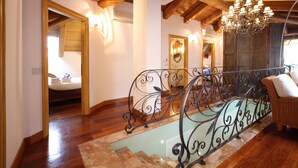 Un escalier en bois avec une rampe en fer forgé, un lustre et un canapé en osier dans une pièce bien éclairée, dont le plafond est orné de poutres en bois.