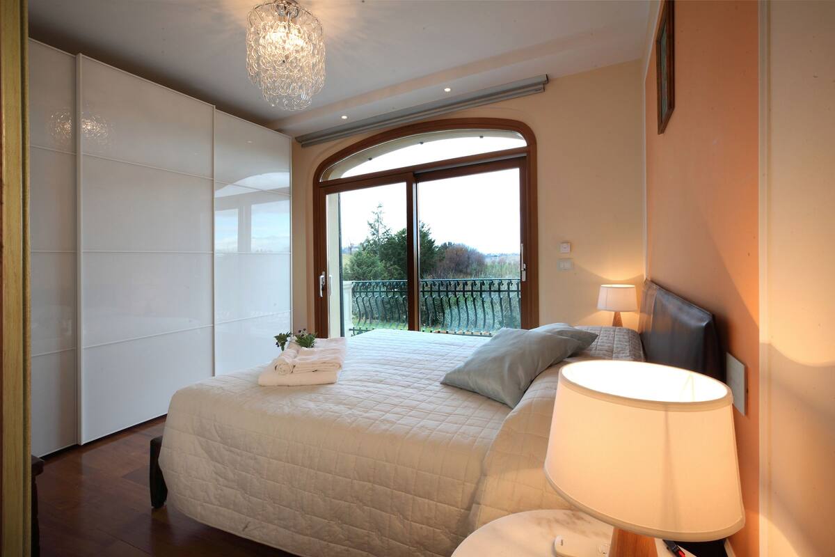 Une chambre à coucher avec un grand lit, une table de chevet, une lampe et une vue sur l’extérieur grâce à une porte vitrée.
