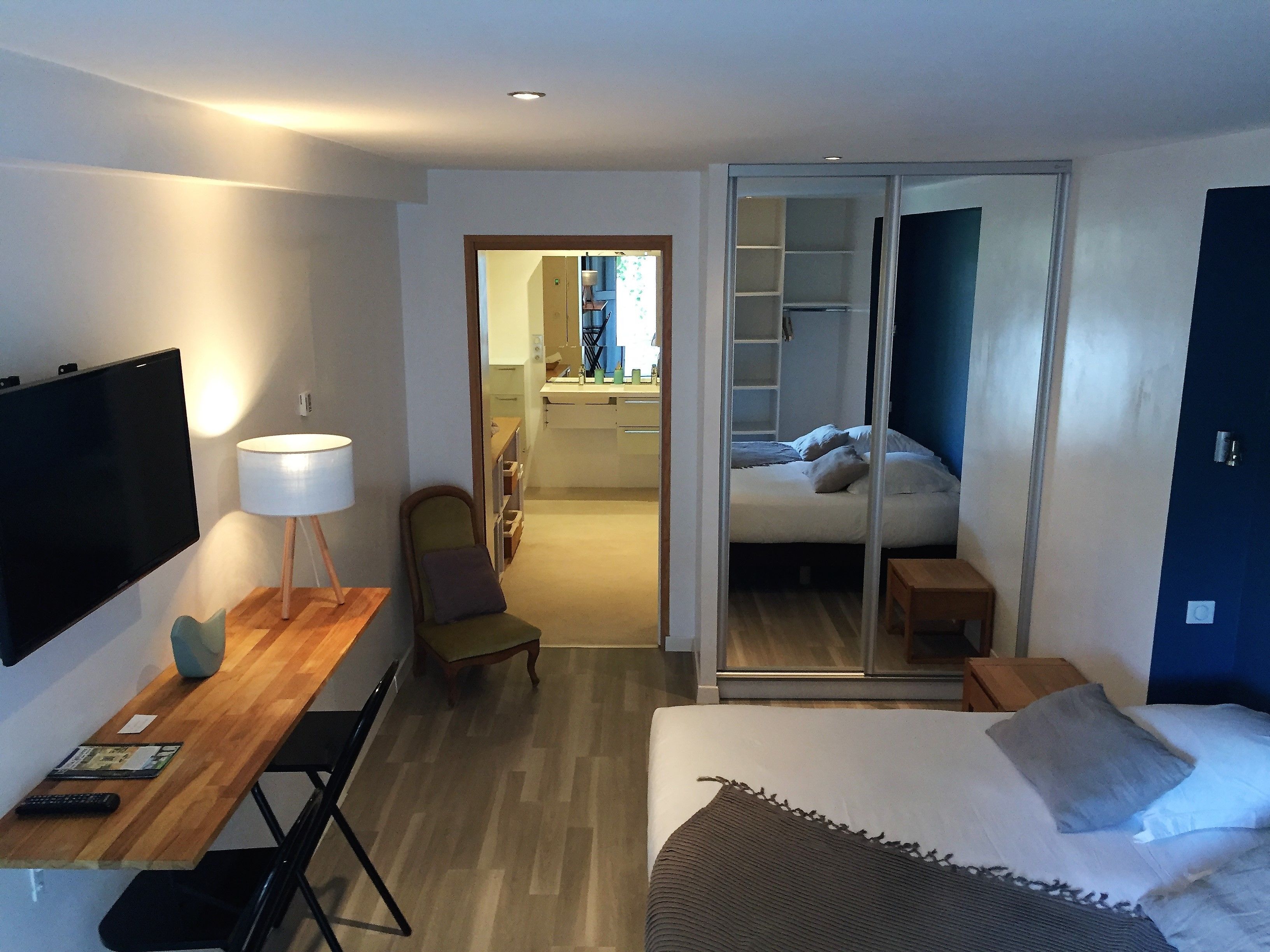 A modern hotel room with a flat-screen TV, a desk with a lamp, a bed with a grey blanket, and a sliding door leading to a bathroom.
