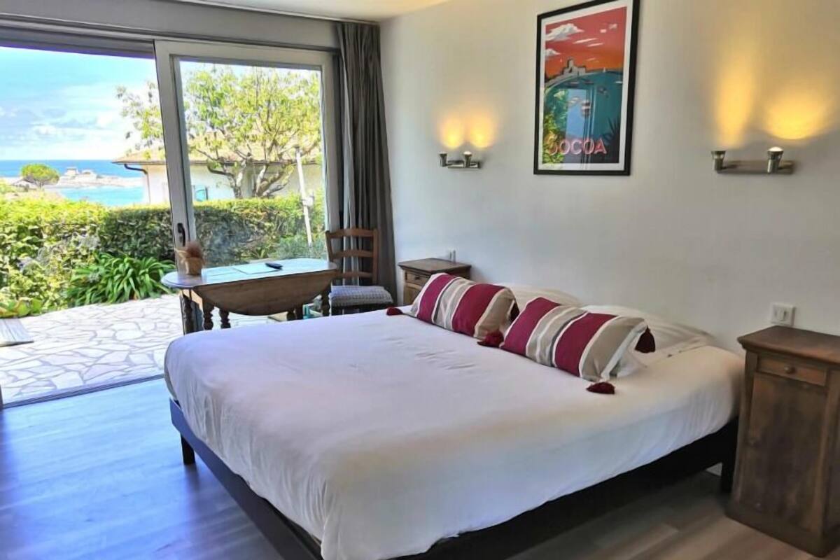 A bedroom with a large bed, a wooden nightstand, a chair, a small table, and a view of the outdoors.