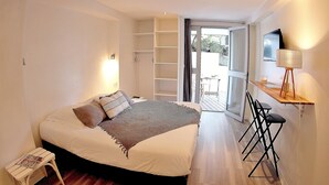 Un dormitorio con cama, una mesita, una silla, un escritorio con lámpara y una puerta corrediza que da a un balcón.