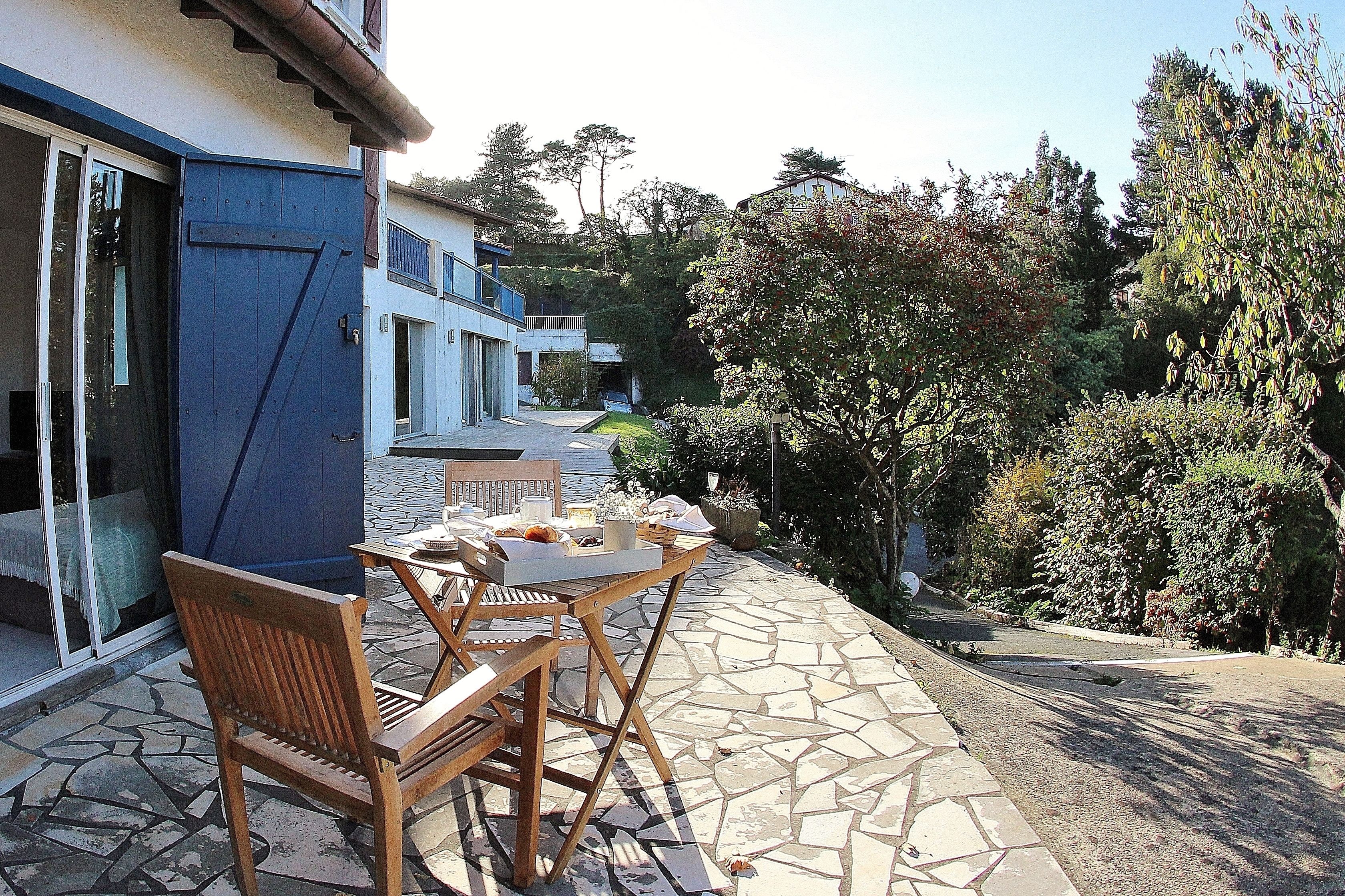 An outdoor patio with a wooden table and chairs, a stone floor, and a view of a residential area with trees and a building in the background.