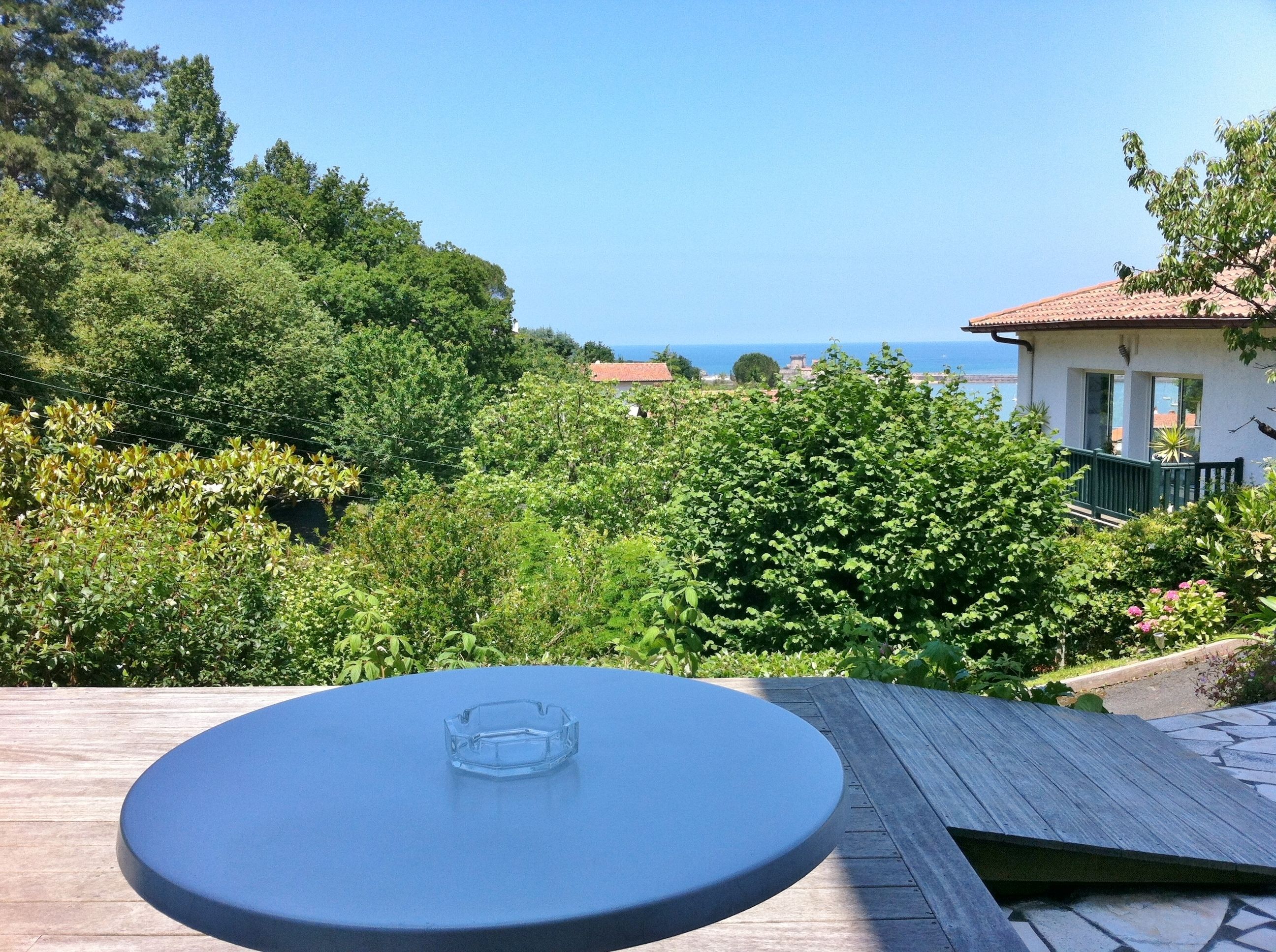 A round table with a clear ashtray on a wooden deck overlooking a lush garden and a coastal view.