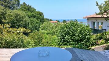 A round table with a clear ashtray on a wooden deck overlooking a lush garden and a coastal view.