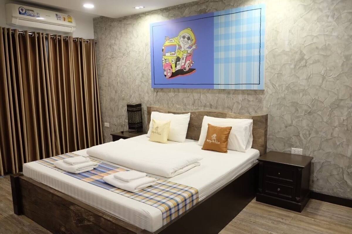 Una cama bien hecha con sábanas blancas y una manta escocesa, una mesita de noche con una lámpara, y una obra de arte en la pared de un personaje de caricatura.
