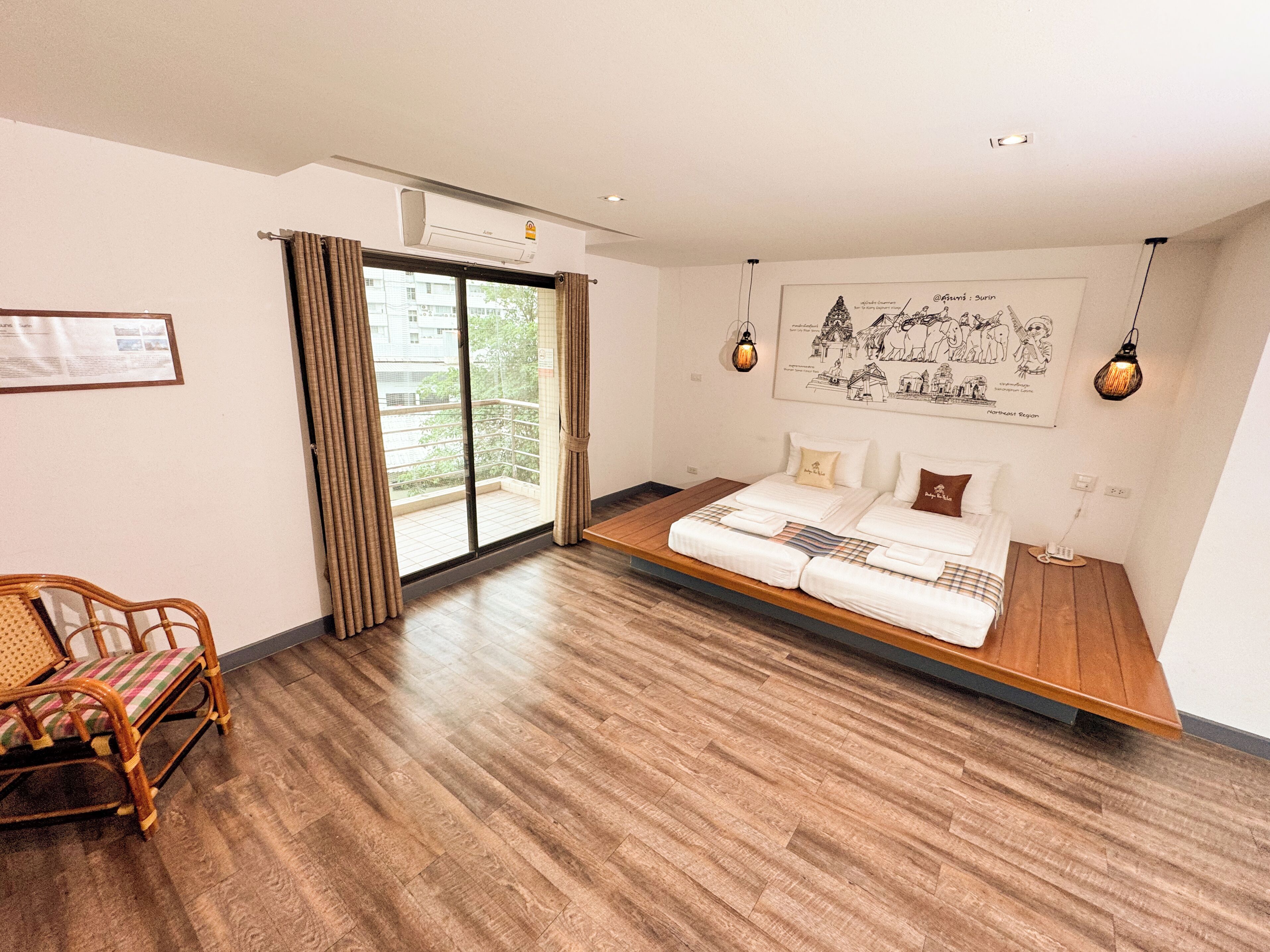 A modern hotel room with a large bed, wooden flooring, a balcony with curtains, a wicker chair, and a wall-mounted light fixture.