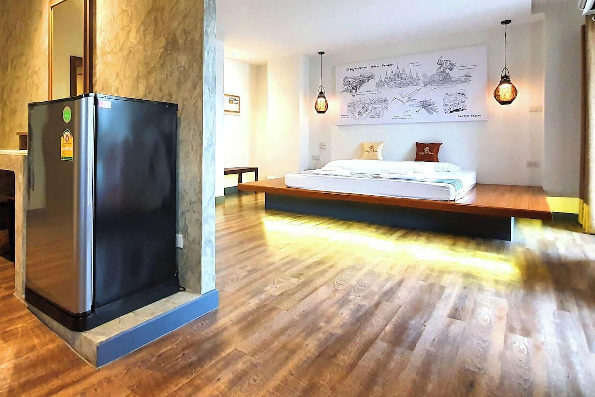 Una habitación de hotel moderna con suelo de madera, una cama con colchón blanco, una mesita pequeña y un refrigerador.