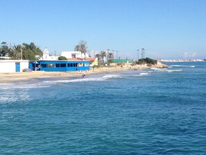 A coastal scene with a blue building, a sandy beach, and the ocean. - Appartement Nabeul Plage (Nabeul)