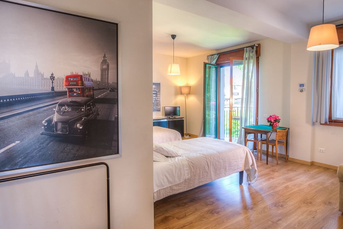 A hotel room with a bed, a desk with a lamp, a table with flowers, and a painting of a cityscape.
