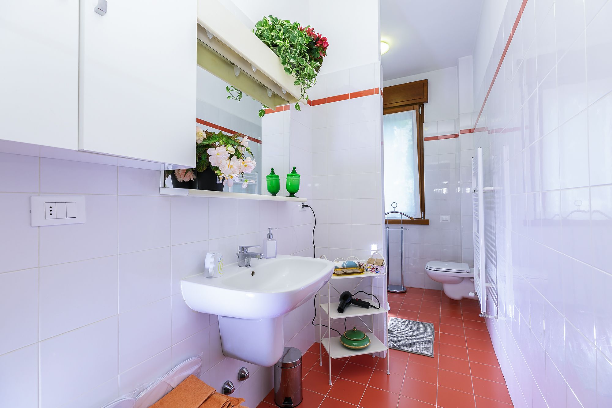 Un baño moderno con un lavamanos blanco, espejo y retrete. Hay una zona de ducha con puerta de cristal y un estante pequeño con botellas verdes y una planta en maceta.