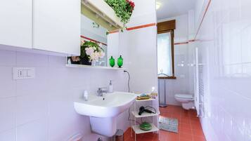 Un baño moderno con un lavamanos blanco, espejo y retrete. Hay una zona de ducha con puerta de cristal y un estante pequeño con botellas verdes y una planta en maceta.